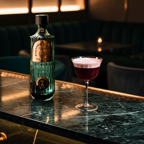 A bottle of Strange Nature gin stands on a dark marble bar counter next to a coupe glass filled with a deep pink cocktail, with a softly blurred bar interior in the background.