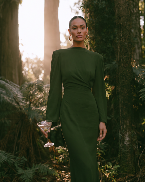 A woman stands in a sunlit forest wearing a fitted long-sleeve dark green midi dress and holding a martini glass.