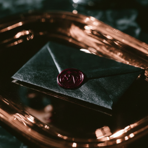 A dark green velvet envelope sealed with a round red wax stamp, resting on a reflective copper tray over a dark marble surface.